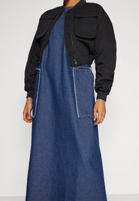 Chaqueta bomber negra corta con dos bolsillos frontales sobre un vestido de denim largo con bordes de bolsillo deshilachados y una textura suave.