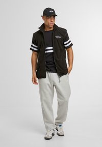 Gilet en polaire noir avec zip, rayures blanches sur les manches, t-shirt noir en dessous, pantalon de survêtement gris, et baskets blanches avec des accents sombres.