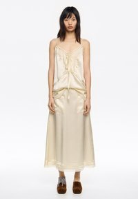 Robe en satin de couleur crème pâle avec des bretelles fines, des détails froncés et des accents en dentelle ; portée avec des chaussures à talons carrés marron ornées de décorations.