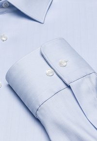 Lichtblauw overhemd met een herringbonepatroon, voorzien van een spread collar, lange manchetten en twee witte knopen op de manchetten.