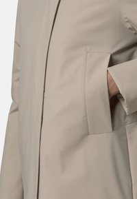 Beżowy wodoodporny płaszcz o prostym kroju, z boczną kieszenią i gładką fakturą. Fason podkreślony przeszyciami z przodu, nadającymi konstrukcję.