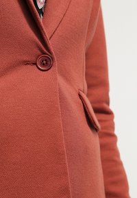 Blazer couleur rouille avec un tissu texturé, une fermeture à un bouton et une poche sur le côté. Le design est élégant et moderne.
