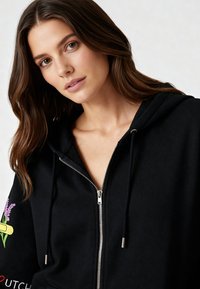 Čierna mikina na zips s veľkým predným vreckom, striebornou kovovou výbavou a farebnými kvetinovými grafikami na rukáve. Mäkká látka a voľný strih.