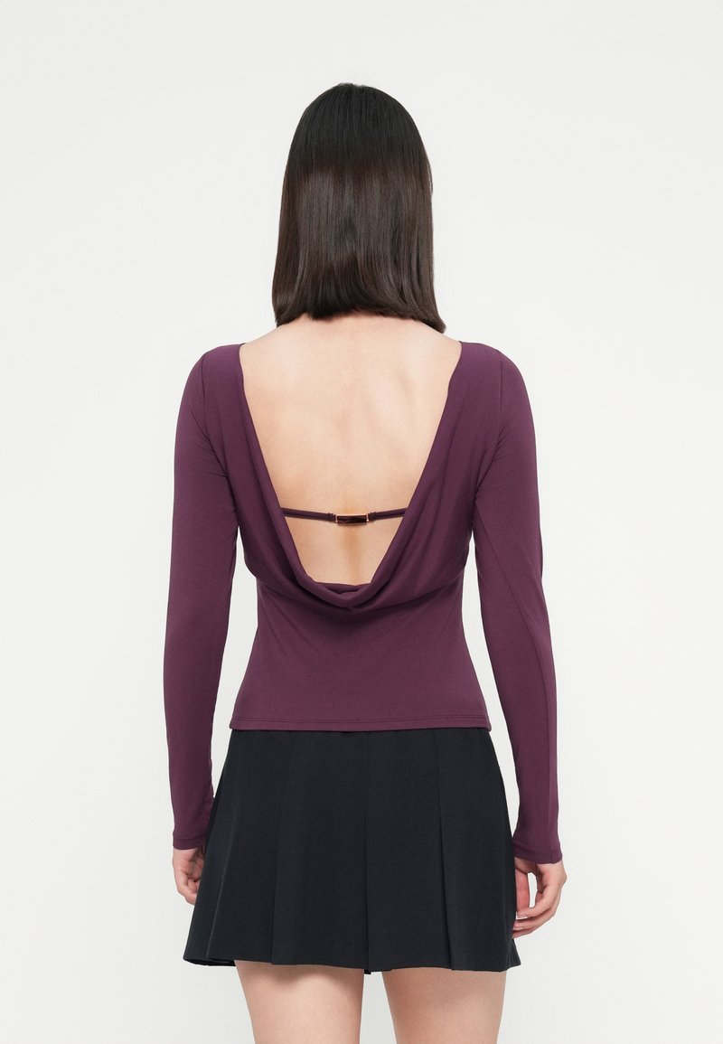 Una donna di spalle indossa una maglietta a maniche lunghe viola con una profonda scollatura aperta sulla schiena e una gonna plissettata nera, su uno sfondo semplice.