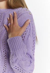 Lila gestrickter Pullover mit Kabelmuster, offenen Maschen und dekorativen silbernen Akzenten an der Schulter. Weicher Stoff und enganliegende Bündchen.