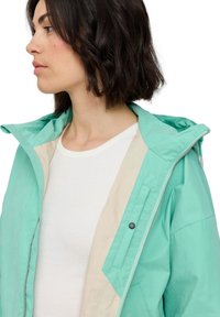 Lightweight jacket in mint green fabric with a contrasting beige interior, featuring a zip closure, hood, and a relaxed fit.