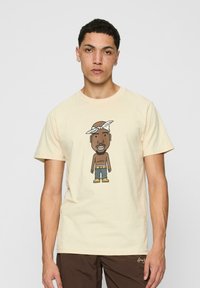 Cream-colored t-shirt featuring a cartoon graphic of a character with a bandana, wearing denim shorts and yellow boots. Short sleeves and crew neck.