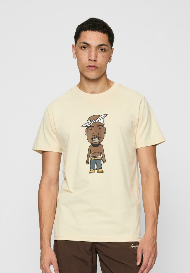 Cream-colored t-shirt featuring a cartoon graphic of a character with a bandana, wearing denim shorts and yellow boots. Short sleeves and crew neck.