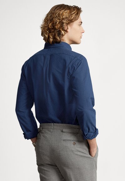 Dark blue button-down shirt with long sleeves, rolled at cuffs, paired with light gray trousers featuring back pockets and a belt.