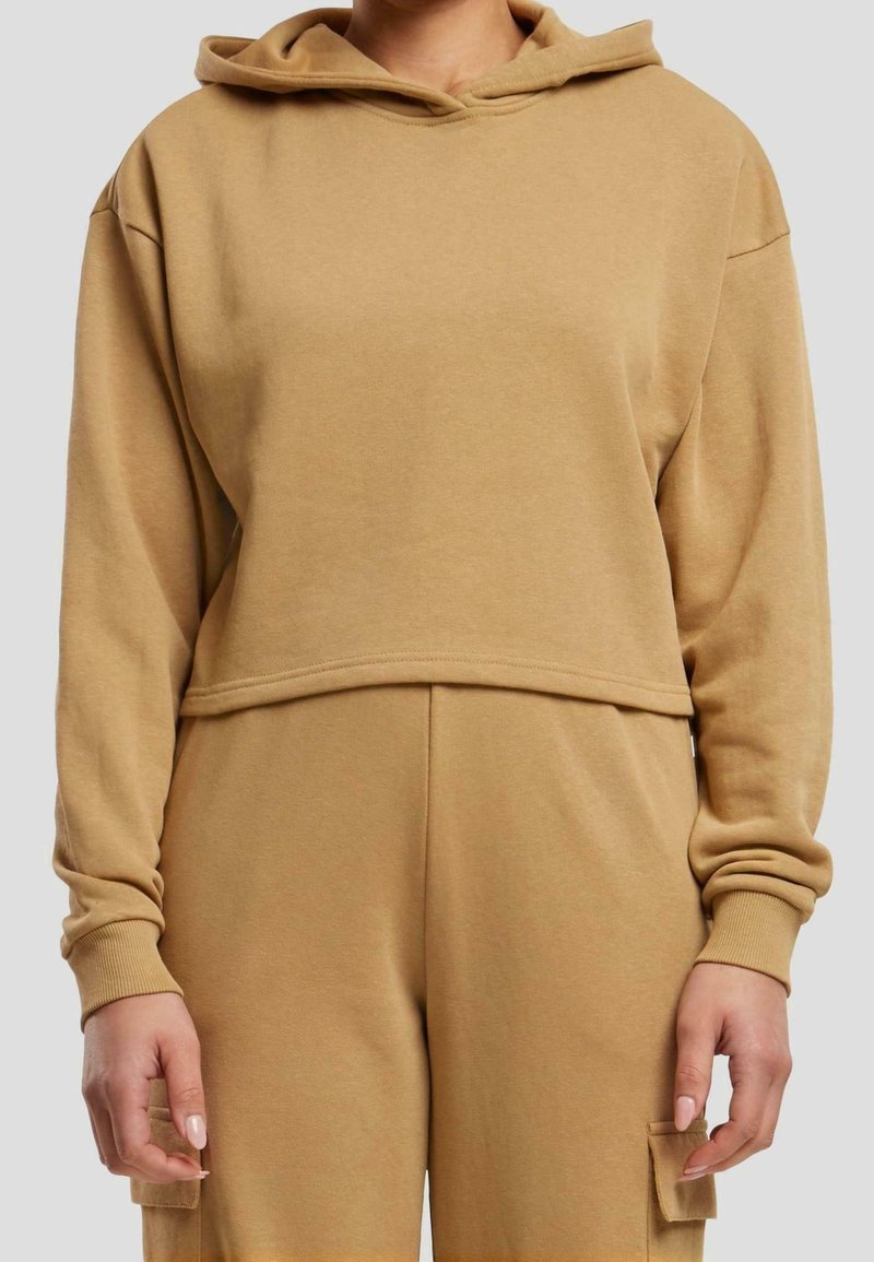 Hoodie beige coupé et pantalon de survêtement taille haute assorti avec poches cargo portés par une personne sur fond uni.