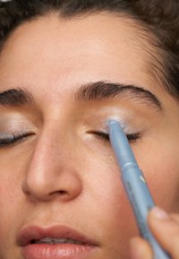 Un crayon à yeux bleu est appliqué sur une paupière avec un fard à paupières argenté scintillant. La peau semble lisse avec un teint naturel.