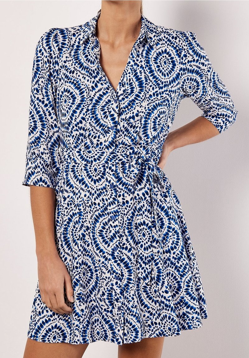 Femme portant une robe portefeuille bleu et blanc à motifs, manches trois-quarts, main sur la hanche, col en V visible.
