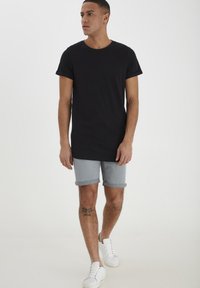 Musta puuvillapaita, harmaat denim-shortsit, joissa on käännetyt lahkeet, valkoiset tennarit ja tatuointi yhdessä jalassa. Malli seisoo valkoista taustaa vasten.
