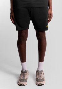 Shorts neri in cotone con logo giallo, dotati di vita elastica e tasche laterali. Indossati con scarpe da ginnastica grigie e calze bianche.