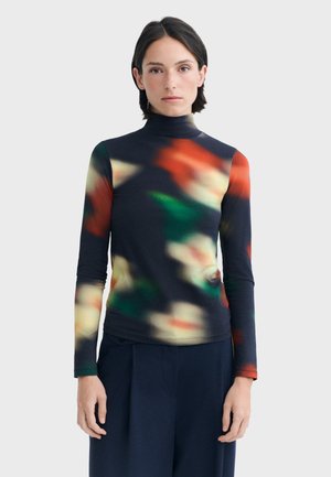Femme aux cheveux courts et foncés portant un pull à col roulé noir avec un motif abstrait flou rouge, vert et crème et un pantalon foncé.