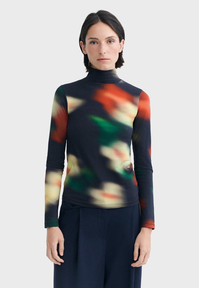 Femme aux cheveux courts et foncés portant un pull à col roulé noir avec un motif abstrait flou rouge, vert et crème et un pantalon foncé.