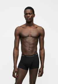 Αθλητικός άνδρας στέκεται φορώντας μαύρα boxer Tommy Hilfiger μπροστά σε απλό ανοιχτό φόντο.