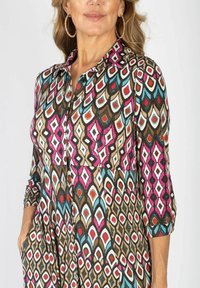 Sukienka z długim rękawem w kolorowy geometryczny wzór w odcieniach różu, czerwieni, niebieskiego i brązu. Posiada kołnierzyk zapinany na guziki oraz kieszenie. Gładka tekstura materiału.