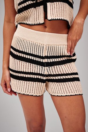 Beige und schwarz gestreifte, gehäkelte Strickshorts, getragen mit einem passenden Crop-Top, das den Bauch zeigt, und zwei an den Seiten ruhenden Händen.