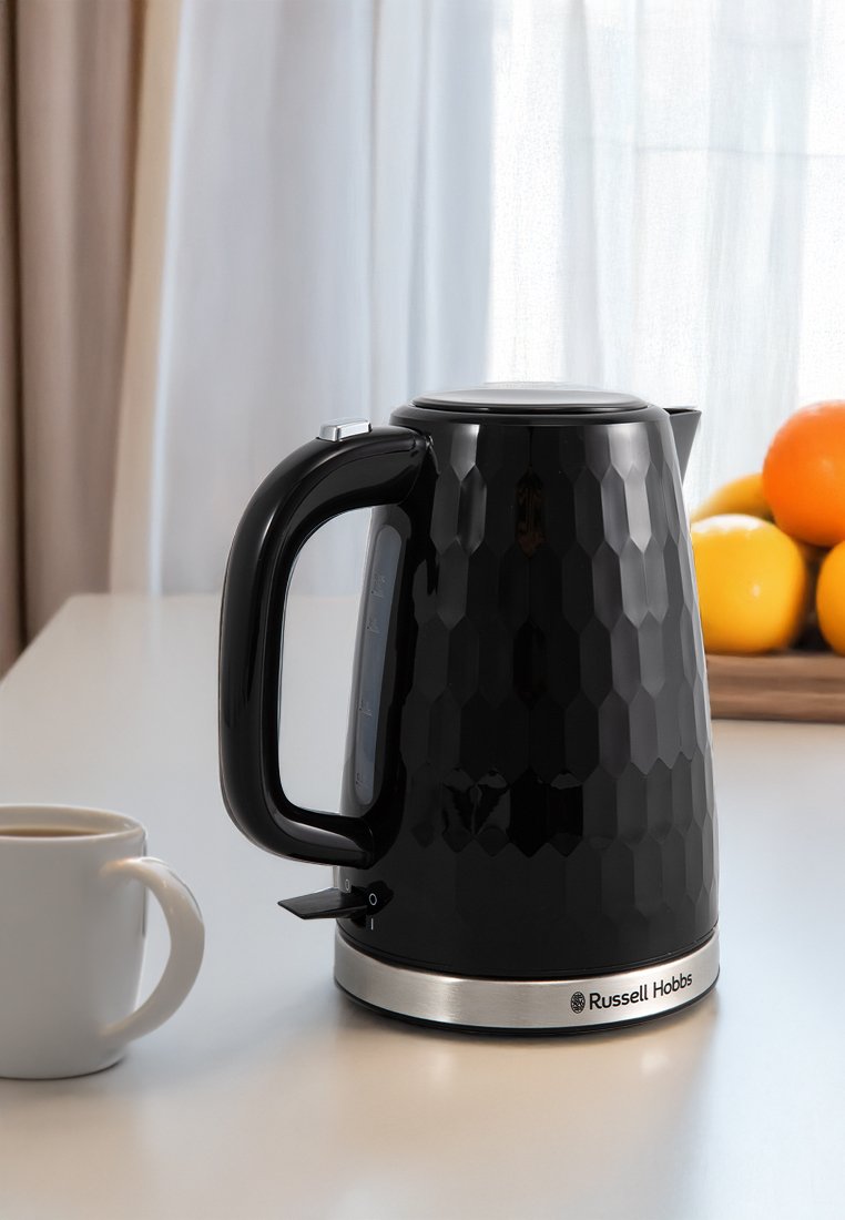 Schwarzer, strukturierter Wasserkocher mit Griff und Ausguss auf weißer Arbeitsplatte, daneben eine weiße Kaffeetasse und ein Tablett mit Orangen im Sonnenlicht.