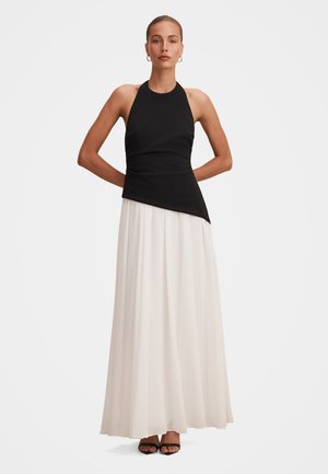 Woman stands with hands behind back wearing sleeveless black asymmetrical top and long flowing white skirt with black open-toe heels.