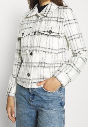 Nainen valkoisessa pörröisessä takissa, jossa musta ruutukuvio ja mustat napit, siniset farkut, käsi osittain taskussa.