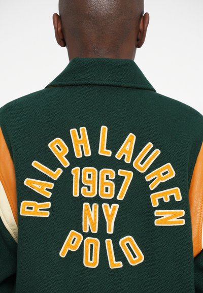 Zaļas vilnas maisījuma jaka ar oranžu ādas plecu akcentu, kuras priekšpusē ir balti izšūti vārdi "RALPH LAUREN 1967 NY POLO."