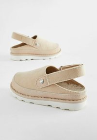 Sabots en velours côtelé beige avec une sangle à l'arrière, semelle en caoutchouc blanc, et un petit accent métallique en forme de chat sur le côté. Doublure intérieure douce visible.