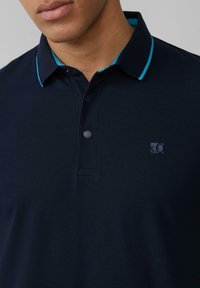 Tumši zilas krāsas polo krekls no strukturētas auduma, ar kontrastējošu gaiši zilu apkakles apdari, trim pogām un mazu logo uz krūtīm.