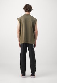 Person with curly hair wearing sleeveless olive green top, black pants with white stripes, and black sneakers with orange accents, standing back.