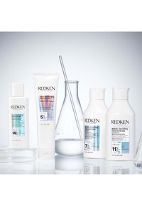 Cuatro botellas de cuidado capilar Redken etiquetadas como concentrado ácido de unión con diferentes porcentajes de ácido cítrico, junto a un vaso de cristal con una varilla de agitación.