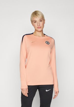 Haut de sport à manches longues en pêche clair avec des accents bleu marine, présentant un col rond, le logo Nike et l'emblème de Chelsea FC sur la poitrine.