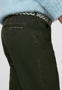 Donkergroene broek met een getextureerde stof, decoratieve stiksels op de zakken en een patroon op de tailleband.