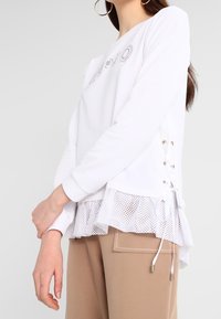 Vit sweatshirt med meshrund kant, snörning i sidorna och strassdetaljer. Långa ärmar och avslappnad passform. Mjukt bomullsmaterial.