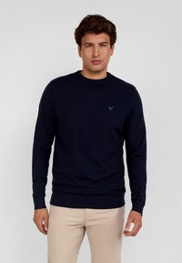 Tamsiai mėlynas megztinis su apvalia iškirpte ir ilgomis rankovėmis, turintis subtilų logotipą ant krūtinės bei raukšlėtas rankogalius ir apačią.