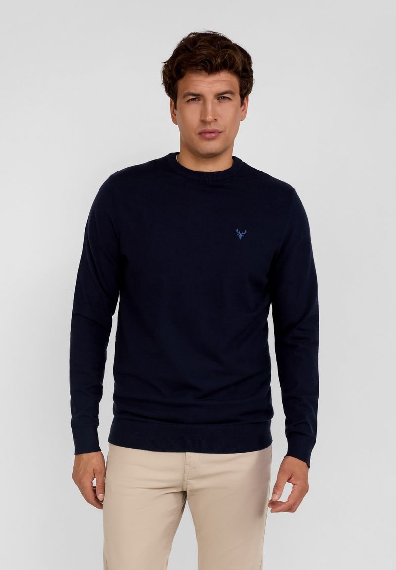 Tamsiai mėlynas megztinis su apvalia iškirpte ir ilgomis rankovėmis, turintis subtilų logotipą ant krūtinės bei raukšlėtas rankogalius ir apačią.