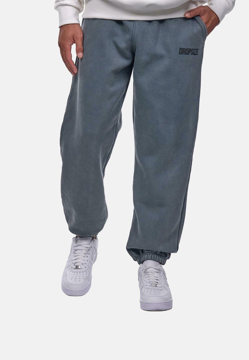 Mørkegrå svedbukser med elastisk talje og manchetter, prydet med et diskret "DROPSIZE" logo på låret. Parret med hvide sneakers.