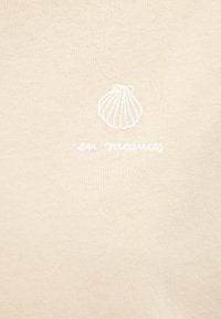 Beige cotton fabric features an embroidered scallop shell design above the text "en tracés" in white. Soft texture and simple design.