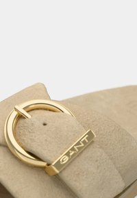 Μπεζ σουέντ σανδάλι με χρυσό κυκλικό πόμολο και μεταλλικό λογότυπο, με λεία, υφή επιφάνεια.
