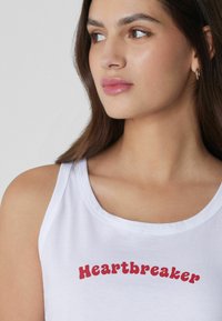 Camiseta sin mangas blanca con el texto rojo "Heartbreaker", con un escote redondo y un diseño simple, hecha de una tela suave.