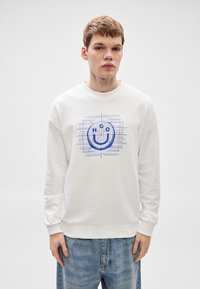 Λευκή φούτερ με μπλε γραφικό σχέδιο που απεικονίζει ένα χαμογελαστό πρόσωπο και το κείμενο "HGO" κεντραρισμένο στο μπροστινό μέρος. Μαλακό ύφασμα, άνετη εφαρμογή.