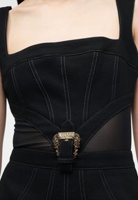 Μαύρο τοπ από τζιν ύφασμα σε στυλ κορσέ με λευκές ραφές, φαρδιές τιράντες, διαφανείς πλαϊνές φάσες και χρυσή διακοσμητική αγκράφα Versace Jeans Couture στη μέση.