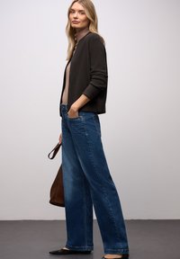 Tmavý kardigan přes béžové tričko, doplněný širokými modrými džínami. Hnědá kabelka, černé baleríny a dlouhé rovné blond vlasy.