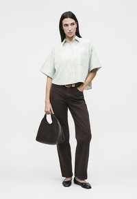 Γυναίκα που φοράει άνετο ριγέ πουκάμισο με κοντά μανίκια, σκουρόχρωμο ίσιο παντελόνι, μαύρες μπαλαρίνες, κρατώντας μια μαύρη τσάντα με το ένα χέρι στην τσέπη.