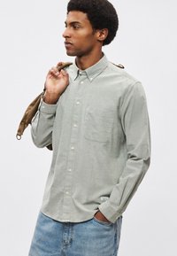 Svetlo zelena srajca z dolgimi rokavi iz mehke tkanine, z ovratnikom na gumbe, enim žepom na prsih in belimi gumbi. Oblečena z modrimi kavbojkami.