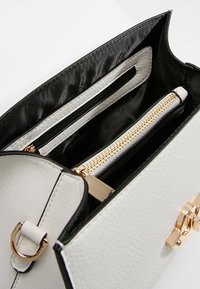 Valkoinen tekstiilinen käsilaukun sisus mustalla vuorella, jossa on kultainen vetoketjupussi ja useita osastoja järjestämistä varten.