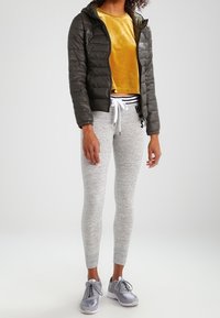 Zateplená tmavě zelená bunda s kapucí, žlutý sametový cropped top, šedé melírové legíny a stříbrné sportovní boty s černými akcenty.