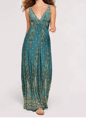 Frau trägt ein türkisfarbenes Maxikleid mit tiefem V-Ausschnitt und aufwendigen gold- und grünen Paisley-Mustern, kombiniert mit weißen Sandalen.
