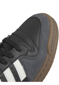 Grå läder- och mockasneaker med svarta detaljer, vita ränder, perforerad tå och en brun gummisul. Har svarta snören och sömmar.