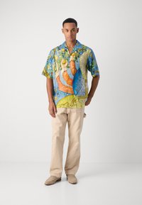 Camisa de manga corta, con un colorido estampado que presenta diseños tropicales en azul, verde y naranja, llevada con pantalones beiges y zapatos claros.